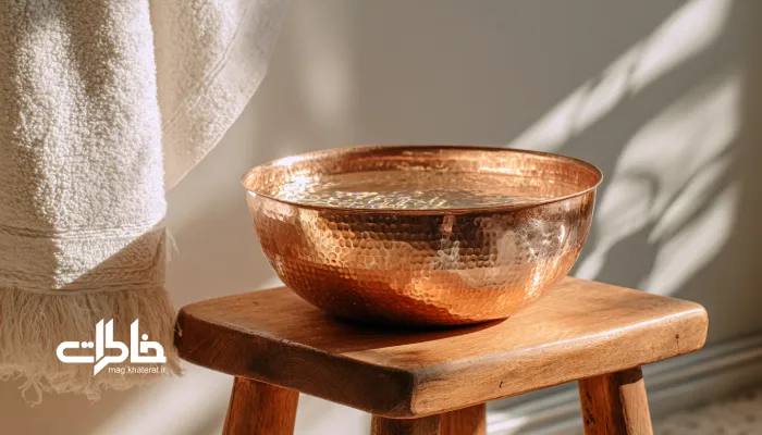 کاسهٔ مسی پر از آب کنار حولهٔ سفید آویزان؛ حس تمیزی و رسم‌های قدیمی. شستن دست و صورت