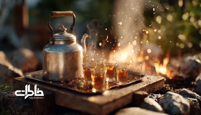 کتری استیل روی آتش در طبیعت، با سینی چای و لیوانهای بخارآلود کنار آن. چای و آتش