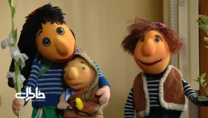 پرتره سینمایی از عروسک الهام‌گرفته از کلاه‌قرمزی با کلاه قرمز، پیراهن آبی‌وسفید راه‌راه، شلوار سبز و نگاه کودکانه در دکور تلویزیونی ایرانی