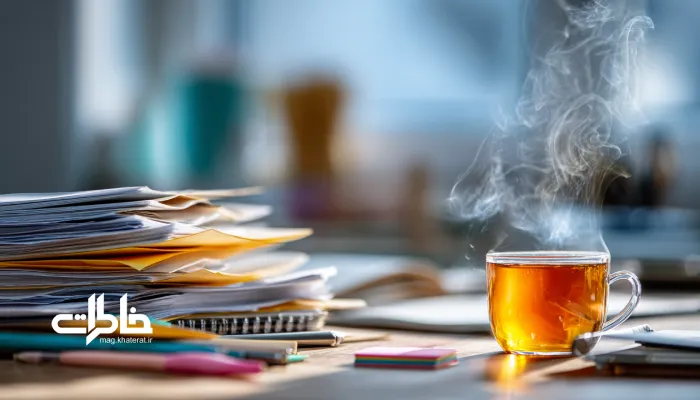 کلوزآپ میز کار شلوغ با پرونده‌ها، دفتر و فنجان چای که نماد آرامش و همدمی در کار است. چای و کار