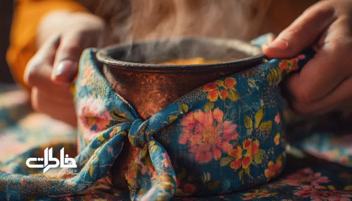 نمای نزدیک از قابلمه غذای خانگی ایرانی که در پارچه بقچه گلدار پیچیده میشود، با بخار غذا و نور گرم طبیعی