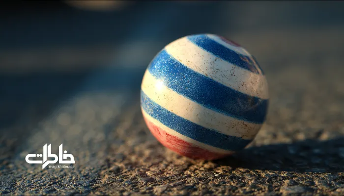 نمای نزدیک از توپ پلاستیکی راه راه سبک و نازک با راه‌راه‌های ریز سفید و آبی، براق و نیمه‌شفاف روی زمین کوچه ایرانی