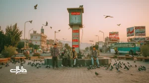 غروب فلکه ساعت با نوجوان‌هایی در پیراهن چهارخانه که کنار برج ساعت عکس یادگاری می‌گیرند؛ اتوبوس، مغازه‌های اطراف و حال‌وهوای نوستالژیک شهری ایران.مجله خاطرات