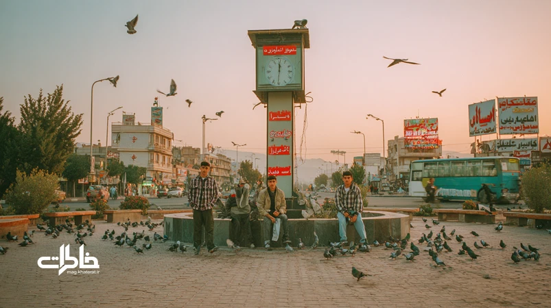 غروب فلکه ساعت با نوجوان‌هایی در پیراهن چهارخانه که کنار برج ساعت عکس یادگاری می‌گیرند؛ اتوبوس، مغازه‌های اطراف و حال‌وهوای نوستالژیک شهری ایران.مجله خاطرات