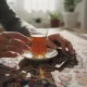 دست‌های ایرانی در خانه امروزی در حال لمس استکان چای، کلید قدیمی و قالی؛ نمادی از اشیای روزمره به عنوان دستگاه زمان در حافظه جمعی