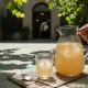 پارچ و لیوان شیشه‌ای شربت خاکشیر با یخ در حیاط ایرانی زیر سایه، یادآور فرهنگ شربت‌های خانگی و پذیرایی تابستانی